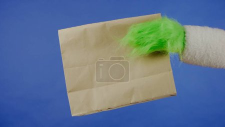 Téléchargez les photos : Grinchs main aux cheveux verts tient un sac en papier sur un fond bleu isolé. Cadeau kidnappeur cosplay. Noel et Nouvel An concept de célébration. Écran bleu, clé chromatique - en image libre de droit