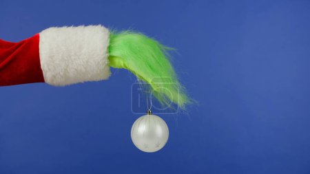 Foto de La mano de pelo verde Grinchs sostiene una bola blanca de Navidad sobre un fondo azul aislado. Cosplay secuestrador de regalos. Concepto de celebración de Navidad y Año Nuevo. Pantalla azul, croma key. - Imagen libre de derechos