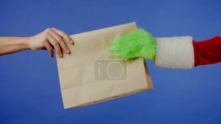Photo for The Grinchs green haired hand reaches out and a mans hand takes a paper bag on an isolated blue background. Gift kidnapper cosplay. Christmas and New Year celebration concept. Chroma, key. - Royalty Free Image