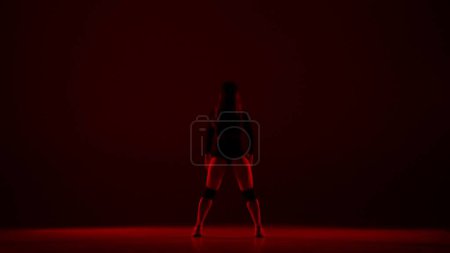 Foto de En el marco sobre un fondo oscuro, se encuentra una mujer joven en ella brilla una luz roja se encuentra una mujer. Demuestra un movimiento de danza al estilo de twerk. Ella es sexy, rítmica. Lleva ropa abierta. - Imagen libre de derechos