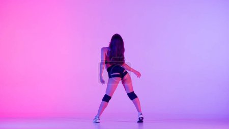 En el marco sobre un fondo multicolor, gradiente se encuentra una mujer joven. Demuestra un movimiento de danza al estilo de twerk. Está de espaldas a la cámara, sexy. Lleva ropa abierta.