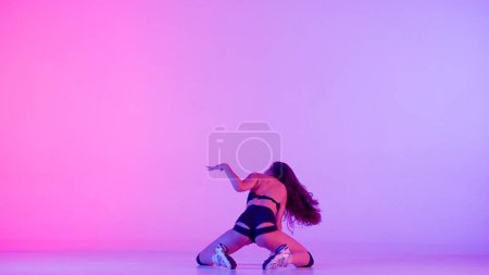 Photo for In frame on multicolored background, gradient kneeling woman. Demonstrates a dance movement in the style of twerk. She stands with her back to the camera, sexy, rhythmic. She is wearing open clothes. - Royalty Free Image