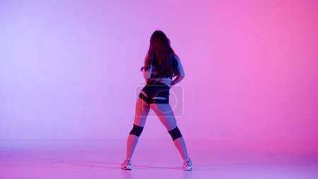 En el marco sobre un fondo multicolor, gradiente se encuentra mujer. Demuestra un movimiento de danza al estilo de twerk. Está de espaldas a la cámara, sexy. Lleva ropa abierta..