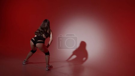 Foto de El marco muestra a una mujer joven sobre un fondo rojo, el centro de atención brilla sobre ella, su sombra es visible. Demuestra un movimiento de danza al estilo de twerk. Ella está vestida con ropa abierta, sexy, rítmica. - Imagen libre de derechos