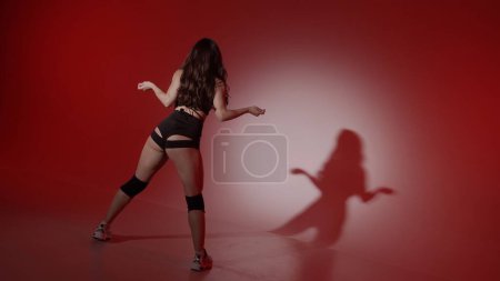 Foto de El marco muestra a una mujer joven sobre un fondo rojo, el centro de atención brilla sobre ella, su sombra es visible. Demuestra un movimiento de danza al estilo de twerk. Ella está vestida con ropa abierta, sexy, rítmica. - Imagen libre de derechos