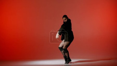 Photo for On a red background. Young woman, showing dance moves towards high heels. A spotlight beam shines on her and gives her a shadow, the edges of the background are darkened. She is sexy, rhythmic. - Royalty Free Image