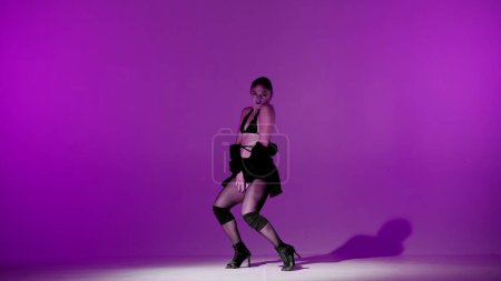 Photo for On purple background. Young woman, showing dance moves towards high heels. A spotlight beam shines on her and gives her a shadow, the edges of the background are darkened. She is sexy, rhythmic. - Royalty Free Image