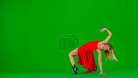 Foto de Enmarcado en un fondo verde, Cromakey. Mujer joven y delgada de pie en vestido rojo. Demuestra un movimiento de baile en la dirección de los tacones altos que descienden al suelo. Ella es sexy, plástico, rítmica - Imagen libre de derechos
