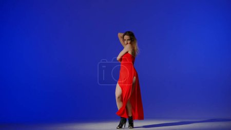 Photo for On a blue background. Young woman, showing dance moves towards high heels. A spotlight beam shines on her and gives her a shadow, the edges of the background are darkened. She is rhythmic, plastic. - Royalty Free Image