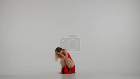 Téléchargez les photos : Encadré sur un fond blanc et gris. Jeune femme, montrant des mouvements de danse vers les talons hauts, accroupi. Elle porte une belle robe rouge et. Elle est sexy, rythmique, en plastique. Plan moyen. - en image libre de droit