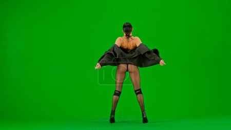 Téléchargez les photos : Encadré sur un fond vert, chromatique. Jeune femme en vêtements révélateurs se tient avec son dos à la caméra. Démontre un mouvement de danse dans la direction des talons hauts. Elle est sexy, rythmique. - en image libre de droit