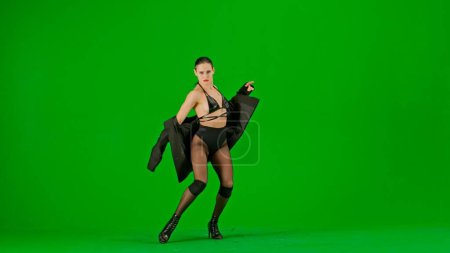 Photo for Framed on a green background, chromakey. Young woman in open dress and jacket, pulling it down lower. Demonstrates a dance move in the direction of high heels. She is sexy, rhythmic. Medium shot. - Royalty Free Image