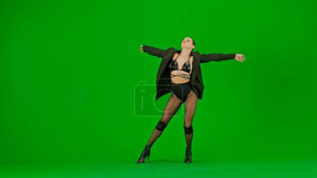 Téléchargez les photos : Encadré sur un fond vert, chromatique. Jeune femme portant des vêtements ouverts et une veste. Démontre un mouvement de danse dans la direction des talons hauts, écartant les bras. Elle est sexy, rythmique. - en image libre de droit