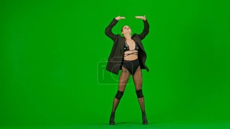 Foto de Enmarcado en un fondo verde, Cromakey. Mujer joven con ropa abierta y chaqueta. Demuestra un movimiento de baile en la dirección de los tacones altos. Es sexy, plástica, rítmica. Tiro medio. - Imagen libre de derechos