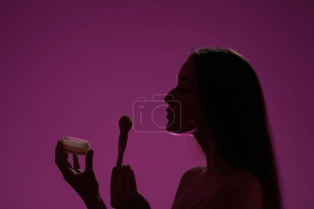 Foto de Vista lateral de una silueta oscura de una mujer sosteniendo una lata de polvo y un cepillo de maquillaje en un estudio sobre un fondo rosa en luz de neón rosa de cerca. Seminude mujer soplando el exceso de polvo de cepillo de maquillaje - Imagen libre de derechos
