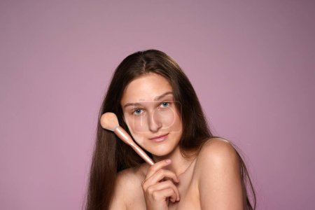 Téléchargez les photos : Portrait d'une jeune femme avec un pinceau de maquillage à la main et des traces de poudre sur le visage. Seminude femme en studio sur fond rose gros plan - en image libre de droit