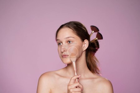 Photo for A woman applying makeup using a makeup brush. Hair of woman pinned up with makeup brushes. Seminude woman in studio on pink background in pink neon light close up. Face contouring makeup - Royalty Free Image