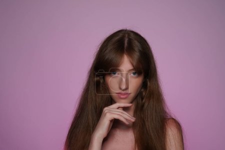 Téléchargez les photos : Portrait de femme aux cheveux longs en studio sur fond rose néon rose clair de près. Femme séminude regardant droit devant. Concept de beauté, cosmétologie, soins - en image libre de droit