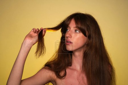 Téléchargez les photos : La jeune femme regarde la mèche de ses cheveux à la main. Séminude femme en studio sur fond jaune dans la lumière jaune néon. Poils fourchus, ternes, secs. Concept de beauté et de soins capillaires - en image libre de droit