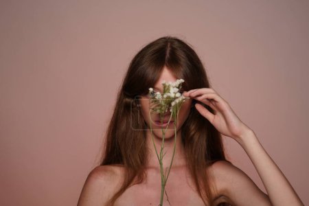 Foto de Joven mujer de pelo largo cubrió su cara con flores silvestres blancas. Mujer Seminude en estudio sobre fondo marrón de cerca. Belleza femenina natural - Imagen libre de derechos