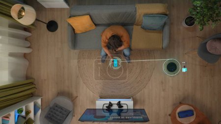 Téléchargez les photos : Smart house technologie concept créatif. Portrait d'homme dans l'appartement. Vue de dessus de l'homme assis sur le canapé dans la chambre avec des gadgets, allumer l'aspirateur. Image numérique infographique sur l'objet. - en image libre de droit