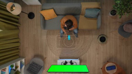 Téléchargez les photos : Smart house technologie concept créatif. Portrait d'homme dans l'appartement. Vue du dessus de l'homme assis sur le canapé tenant smartphone avec application pour gadgets maison, tv avec écran vert chromatique. - en image libre de droit