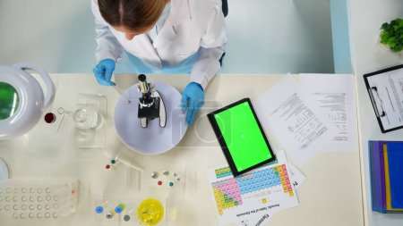 Photo for There is a close up of a woman sitting in the frame above. She is a scientist, analyzing material from a test tube in a microscope. A tablet with a green screen lies nearby. Place for advertising. - Royalty Free Image