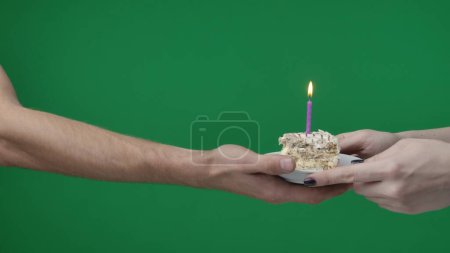 Téléchargez les photos : Gros plan sur fond vert chromatique. Une femme et un homme, vous pouvez voir leurs mains. Elle tient un morceau de gâteau avec une bougie allumée et le remet à l'homme. C'est son anniversaire, elle le félicite. - en image libre de droit