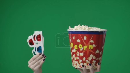 Photo for In the close up shot on the green background. A womans hand that is raised up and holds 3D glasses red blue and a box, a bucket of popcorn. Showing off to a movie theater for a movie - Royalty Free Image