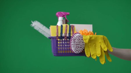 Photo for Framed on a green background. A womans hand that holds out a basket containing detergent, gloves and a brush. Items for cleaning, she holds out a box to the camera. - Royalty Free Image