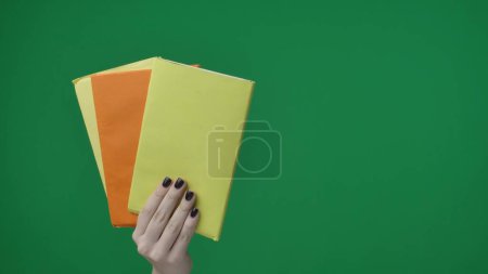 Téléchargez les photos : Dans le cadre sur un fond vert, une main féminine chromakée avec une manucure tient des feuilles de papier lisses et vides, elles sont jaunes et orange. Elle les a tenus et les a montrés à la caméra. Cadre moyen - en image libre de droit