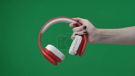 Photo for In the frame on a green background, a lame key female hand with a manicure holds and extends red wireless headphones for listening to music. Here can be your advertising. Medium frame. - Royalty Free Image
