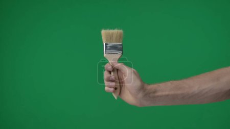 Photo for In the frame on a green background, a chromakey male hand that holds, a tool, a brush on a wooden handle with a natural shield, it is used to paint walls and canvases. Here can be your advertisement. - Royalty Free Image