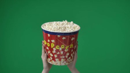 Photo for In the shot close up on the green background. A womans hand that is raised up and holds a box, a bucket of popcorn. Shows her going to a movie theater to see a movie. She holds it over her two hands. - Royalty Free Image