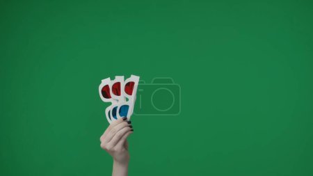 Photo for In the close up shot on the green background. A womans hand that is raised up and holds 3D glasses red blue. She shows the movie at a movie theater at a show with glasses to improve the effect. - Royalty Free Image