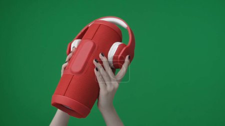 Téléchargez les photos : Dans le cadre sur un fond vert, une main de femme boiteuse avec une manucure tient un haut-parleur rouge portable sur lequel sont placés des écouteurs sans fil. Elle les a élevés et les montre à la caméra. - en image libre de droit