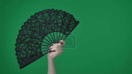 Photo for In the frame on a green background, a chrome key female hand with a manicure holds a black, openwork fan. She opened it, used to inflate the air. Here can be your advertising. Medium frame. - Royalty Free Image