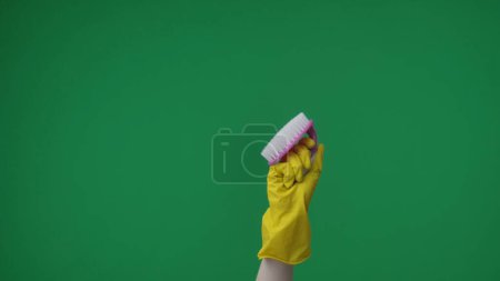 Foto de En el marco sobre un fondo verde, una mano femenina coja con un guante de goma amarillo sostiene y demuestra un cepillo. Se utiliza para la limpieza y artículos de limpieza. Aquí puede estar tu publicidad. Marco medio. - Imagen libre de derechos