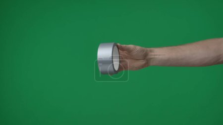 Photo for In frame on green background, chromakey male hand that holds, holds out scotch tape, silver colored tape. It is used for sticking on all surfaces. This could be a place for your advertisement. - Royalty Free Image