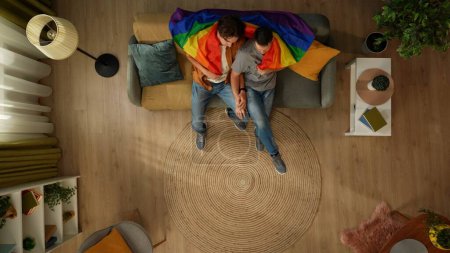 Téléchargez les photos : Vue de dessus prise d'un couple homosexuel à la maison. Ils sont assis sur le canapé enveloppé dans le drapeau LGBTQ, regardant la télévision, choisissant quoi regarder. Égalité, contenu éducatif, fierté. - en image libre de droit