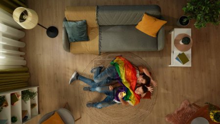 Téléchargez les photos : Vue de dessus prise d'un couple homosexuel couché sur le sol ensemble, embrassant et prenant des selfies sur un smartphone enveloppé dans un drapeau LGBT pour encourager l'amour, l'égalité, la liberté. Contenu éducatif, fierté. - en image libre de droit