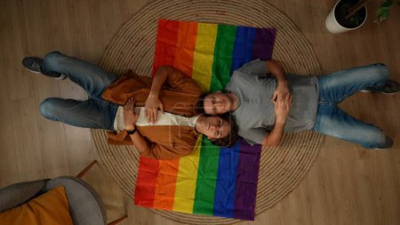Foto de Vista superior de una pareja homosexual, bisexual, pansexual, transgénero que pone en la bandera LGBT, mirando a la cámara para fomentar el amor, la igualdad, la libertad. Contenido educativo, libertad de amor, orgullo. - Imagen libre de derechos