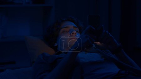 Téléchargez les photos : Un jeune homme envoie des textos sur son smartphone couché dans son lit à la maison la nuit de près. Technologie, internet, communication et concept humain - en image libre de droit