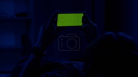 Téléchargez les photos : Un homme allongé sur le canapé la nuit et tenant horizontalement un smartphone avec un écran vert. L'homme regarde une vidéo, un film ou une série. Espace publicitaire, lieu de travail - en image libre de droit