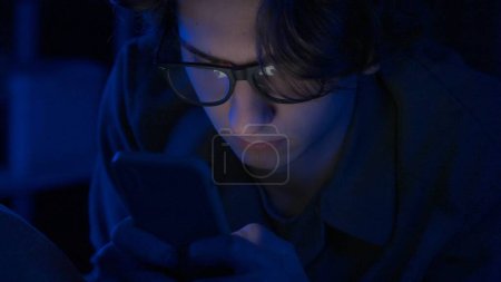 Téléchargez les photos : Un jeune homme portant des lunettes utilise un smartphone couché au lit à la maison la nuit de près. Un homme envoie des SMS, joue à un jeu en ligne ou parcourt des photos, des vidéos. Technologie, internet, communication et - en image libre de droit