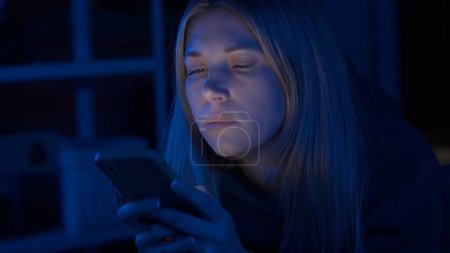 Foto de Mujer atractiva usando teléfono inteligente acostado en la cama en casa por la noche de cerca. Una mujer está enviando mensajes de texto, jugando un juego en línea o navegar por fotos, videos. Tecnología, internet, comunicación y concepto de personas - Imagen libre de derechos