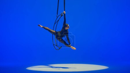 Photo for Rhythmic gymnastics girl performs the scum on one arm in the air on a metal rotating structure moon. Blue background. - Royalty Free Image