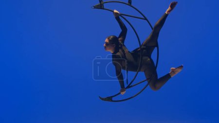 Foto de Chica de gimnasia rítmica realiza la escoria en un brazo en el aire en una luna estructura giratoria de metal. Fondo azul. - Imagen libre de derechos