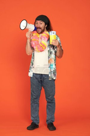 Photo for A man speaks into a megaphone and holds up a passport with airline tickets. A man wearing Hawaiian beads in the studio on orange background full length. Vertical shot. Air flight journey concept - Royalty Free Image