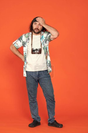 Photo for A male traveler with a retro camera around his neck is thinking anxiously about something, clutching his head with his hand. A man in a colorful shirt, wearing crocs, stands at full height in the - Royalty Free Image